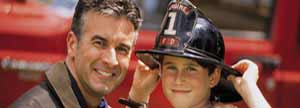 Boy wearing firefighter helmet next to firefighter